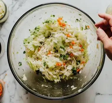 A hand mixing a bowl of chopped vegetables, such as cabbage and carrots, surrounded by various ingredients on a countertop.