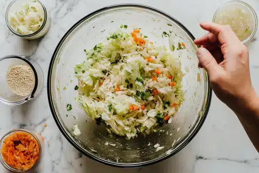 A hand mixing a bowl of chopped vegetables, such as cabbage and carrots, surrounded by various ingredients on a countertop.