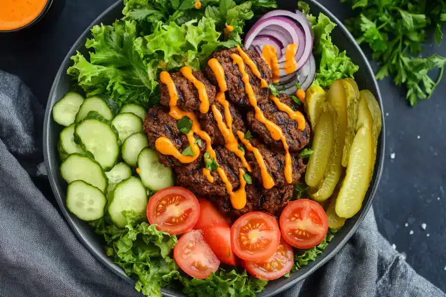 A vibrant salad bowl filled with lettuce, cucumbers, tomatoes, pickles, and seasoned meat drizzled with orange sauce.