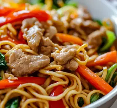 A bowl of flavorful stir-fried noodles with vegetables and chicken pieces.