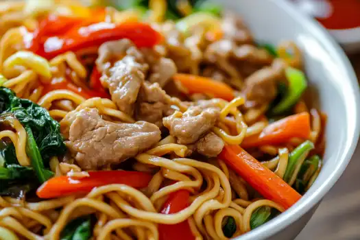 A bowl of flavorful stir-fried noodles with vegetables and chicken pieces.