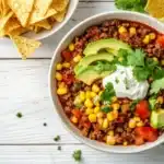 A bowl of chili topped with avocado, sour cream, and cilantro sits on a white wooden table next to tortilla chips.