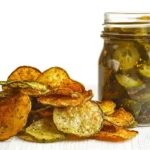 A pile of crispy vegetable chips next to a jar of pickles on a white surface.