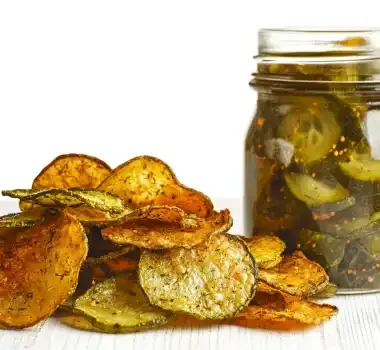 A pile of crispy vegetable chips next to a jar of pickles on a white surface.