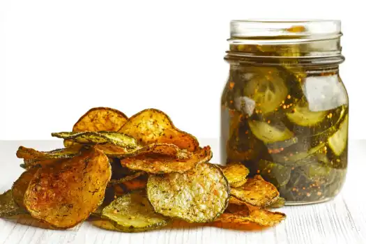 A pile of crispy vegetable chips next to a jar of pickles on a white surface.